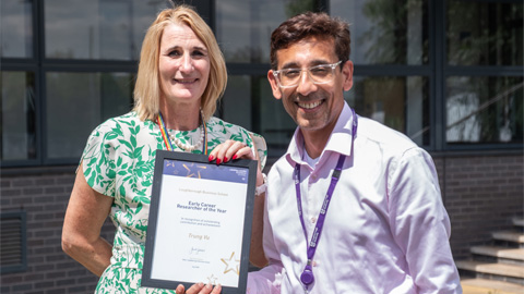 Two people standing outdoors in front of a building with large windows; one holds a framed 